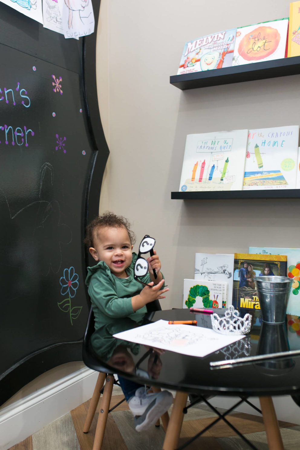 Laughing toddler girl holding crayons while sitting at a child table with coloring material in front of her